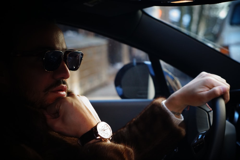 Hombre con gafas de sol negras, reloj de color dorado y chaqueta marrón colocando la mano en la barbilla dentro del automóvil