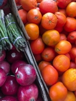 A vibrant assortment of fresh produce including bright orange oranges, deep red pomegranates, and green asparagus bundled together. The fruits and vegetables are displayed in cardboard boxes, creating a colorful and appetizing arrangement.