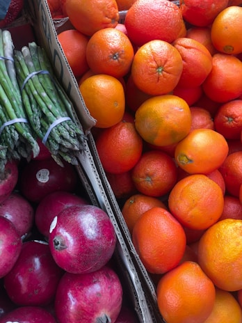 A vibrant assortment of fresh produce including bright orange oranges, deep red pomegranates, and green asparagus bundled together. The fruits and vegetables are displayed in cardboard boxes, creating a colorful and appetizing arrangement.