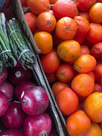 A vibrant assortment of fresh produce including bright orange oranges, deep red pomegranates, and green asparagus bundled together. The fruits and vegetables are displayed in cardboard boxes, creating a colorful and appetizing arrangement.