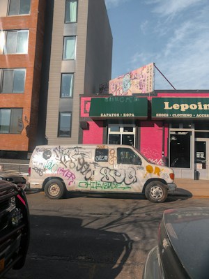 A white van is parked on the street, covered in various graffiti and tags. It's positioned in front of a storefront with a green and pink sign that includes the words 'Zapatos' and 'Ropa', likely indicating a store selling shoes and clothing. The backdrop features multi-story buildings with a clear sky above.