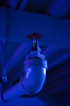 A close-up view of a metallic industrial pipe leading to a sprinkler or nozzle, illuminated by a blue light. The pipe has a shiny surface and connects to a red circular component mounted under a ceiling with a ribbed structure.