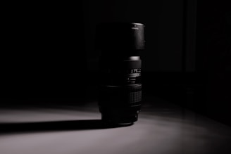 A camera lens stands upright on a reflective surface, casting a long shadow. The scene is dimly lit, creating a dramatic contrast between the dark lens and its surroundings.