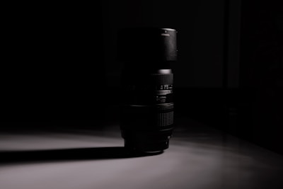 A camera lens stands upright on a reflective surface, casting a long shadow. The scene is dimly lit, creating a dramatic contrast between the dark lens and its surroundings.