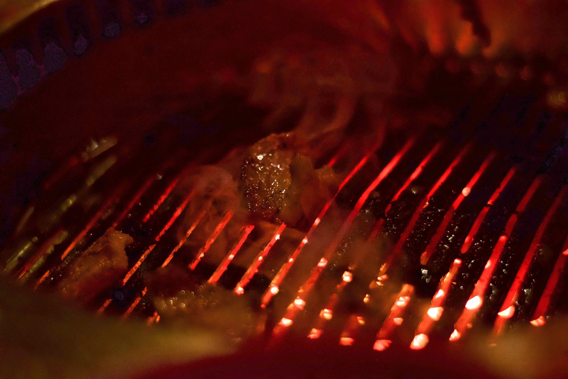 A close-up view of a grill with glowing red coals and pieces of meat being cooked. The grates are illuminated by the intense heat from below, creating a pattern of red and yellow light. Smoke is visible rising from the cooking meat.