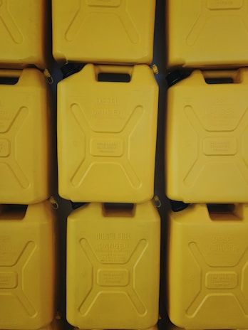 A stack of yellow jerry cans arranged in a symmetrical pattern. Each can has embossed text reading 'Diesel Fuel' and 'Danger,' indicating they are used for storing flammable liquids. The lighting creates subtle shadows between the cans, adding depth to the image.