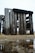 Weathered wooden pallets are stacked together in an open field. The wood appears aged and worn, with variation in texture and color. Below, there is a patch of grass and a small puddle of water reflecting part of the structure.