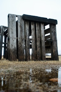 Weathered wooden pallets are stacked together in an open field. The wood appears aged and worn, with variation in texture and color. Below, there is a patch of grass and a small puddle of water reflecting part of the structure.
