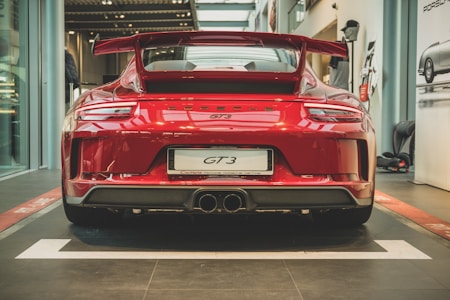 A sleek red sports car with a prominent rear spoiler and dual exhausts displayed in a modern showroom. The car's design emphasizes performance and luxury, evident from its aerodynamic lines and glossy finish.