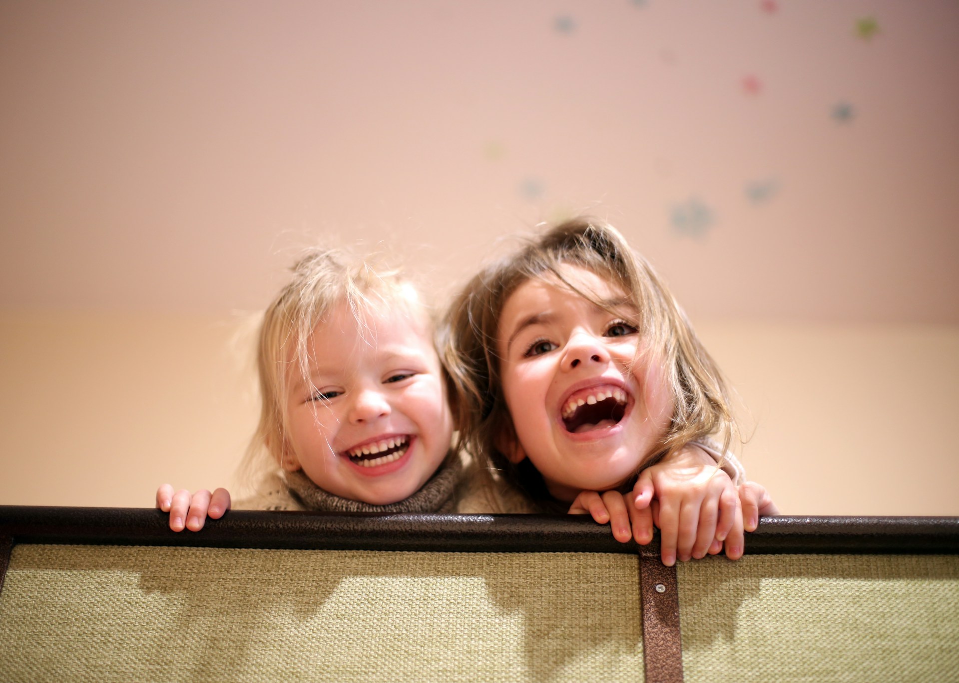 Two children are joyfully smiling and laughing while peering over the edge of a bed frame or railing. The younger child has light blonde hair and is wearing a light-colored sweater, while the older child has darker hair. The background features a soft, neutral color with faint colorful spots, possibly decorations on the ceiling.