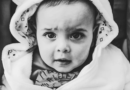 A close-up black-and-white portrait of a baby wrapped in a soft blanket. The baby has large, expressive eyes and is looking directly at the camera. The blanket has a geometric pattern on the inside of the hood.