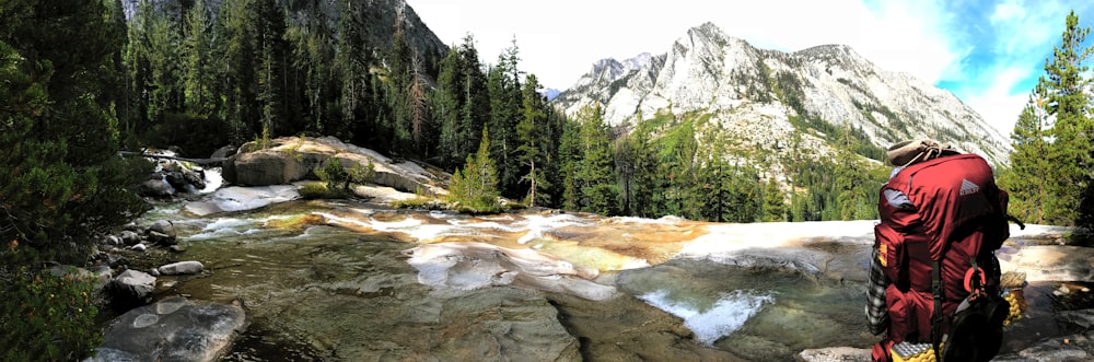 red hiking backpack placed on stone beside river facing trees and mountain