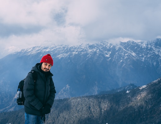 A person wearing a red beanie and a dark winter jacket stands in front of a mountainous landscape with snow-capped peaks and cloudy skies. The person is also carrying a backpack and appears to be in a cold, outdoor setting.