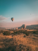 Hot air balloons float serenely over a vast, dry landscape with some greenery and distant hills. The sky is clear with warm hues, indicating a sunrise or sunset.
