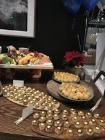 An assortment of finger foods is displayed on wooden trays, featuring small pastries with white toppings and a platter of golden-brown puff pastries. A vibrant fruit platter, including watermelon, pineapple, strawberries, and blueberries, is accompanied by a poinsettia plant in the background. Blue balloons are visible in the upper right corner, suggesting a festive or celebratory setting.