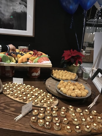 An assortment of finger foods is displayed on wooden trays, featuring small pastries with white toppings and a platter of golden-brown puff pastries. A vibrant fruit platter, including watermelon, pineapple, strawberries, and blueberries, is accompanied by a poinsettia plant in the background. Blue balloons are visible in the upper right corner, suggesting a festive or celebratory setting.