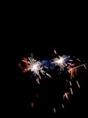 Two sparklers are held in a hand against a dark background, emitting bright sparks and small flares of light.