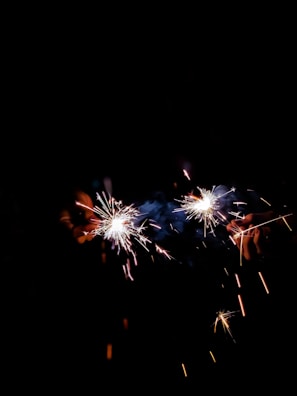 Two sparklers are held in a hand against a dark background, emitting bright sparks and small flares of light.