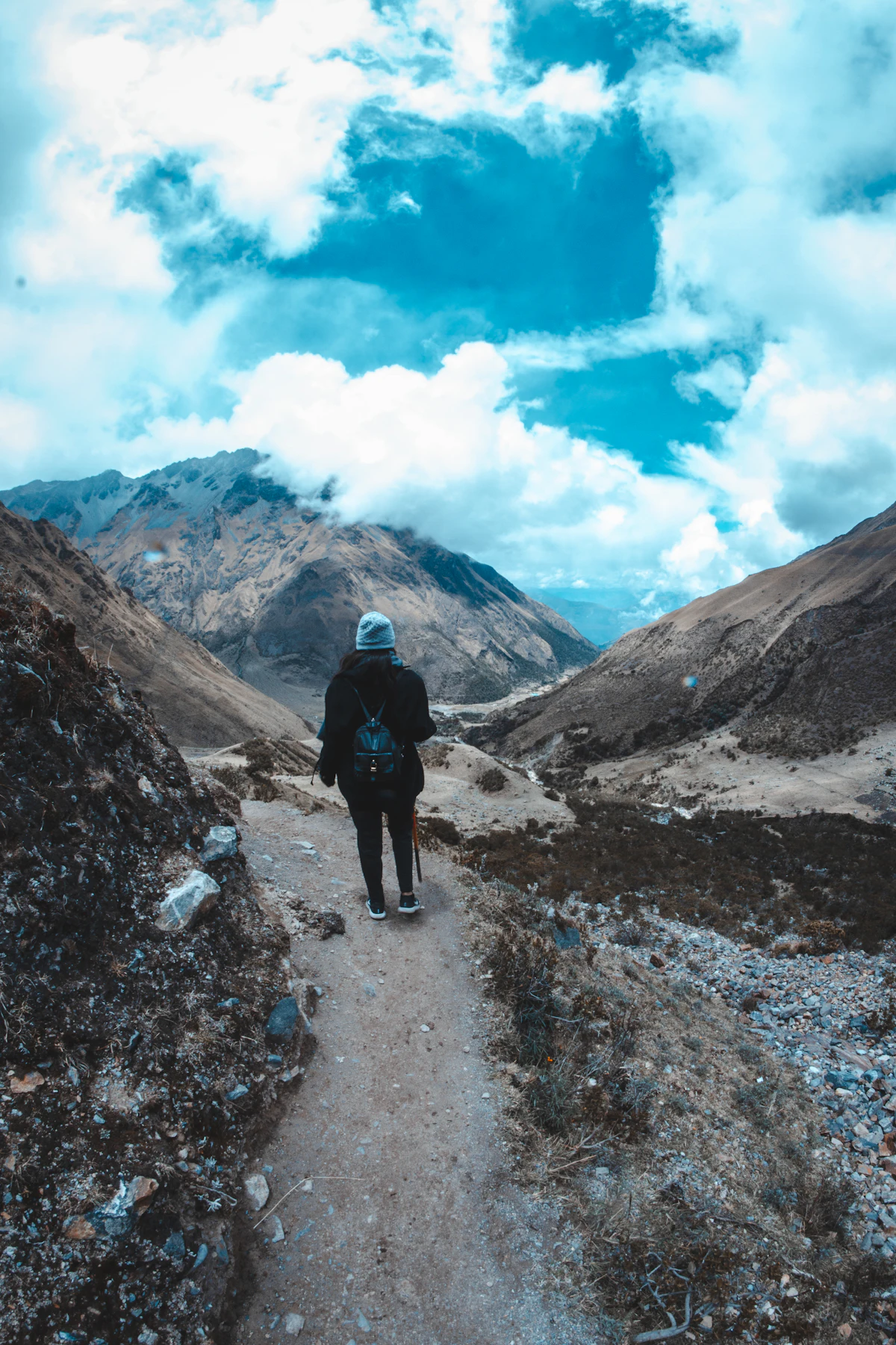 A hiker in dark clothing standing on a mountain trail overlooking a vast valley and peaks