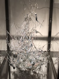 An intricate sculpture composed of twisted metal wires adorned with small, reflective beads and discs. The structure resembles an abstract, organic form with various branches extending upwards. It is enclosed within a transparent display case, creating reflections and shadows on the surrounding surfaces.