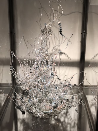 An intricate sculpture composed of twisted metal wires adorned with small, reflective beads and discs. The structure resembles an abstract, organic form with various branches extending upwards. It is enclosed within a transparent display case, creating reflections and shadows on the surrounding surfaces.