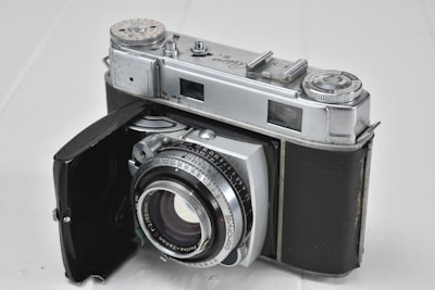 A vintage film camera with a silver metallic body and black leather covering. It has a classic design featuring manual dials and a lens with aperture and focus markings. The camera's door is open, revealing the lens and internal components.