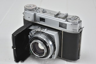 A vintage film camera with a silver metallic body and black leather covering. It has a classic design featuring manual dials and a lens with aperture and focus markings. The camera's door is open, revealing the lens and internal components.