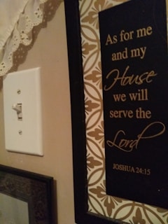 A wall with a decorative plaque featuring the text 'As for me and my House we will serve the Lord Joshua 24:15'. The plaque has a black background with elegant gold script surrounded by a patterned border. To the left, there is a white light switch and some lace fabric at the top corner.