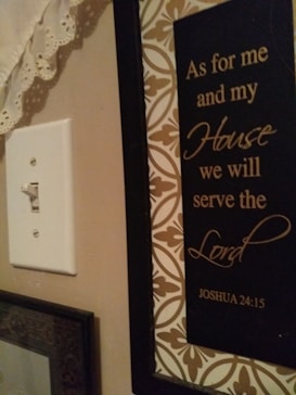 A wall with a decorative plaque featuring the text 'As for me and my House we will serve the Lord Joshua 24:15'. The plaque has a black background with elegant gold script surrounded by a patterned border. To the left, there is a white light switch and some lace fabric at the top corner.