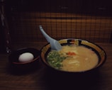 A bowl of creamy ramen soup is accompanied by a white egg in a separate black bowl. The ramen is topped with green onions and a hint of red seasoning, with a blue spoon resting inside the bowl. The bowls are set on a wooden surface with a backdrop of bamboo paneling, creating a warm, cozy atmosphere.