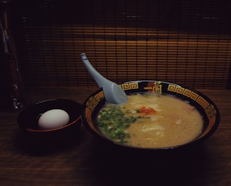 A bowl of creamy ramen soup is accompanied by a white egg in a separate black bowl. The ramen is topped with green onions and a hint of red seasoning, with a blue spoon resting inside the bowl. The bowls are set on a wooden surface with a backdrop of bamboo paneling, creating a warm, cozy atmosphere.