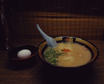 A bowl of creamy ramen soup is accompanied by a white egg in a separate black bowl. The ramen is topped with green onions and a hint of red seasoning, with a blue spoon resting inside the bowl. The bowls are set on a wooden surface with a backdrop of bamboo paneling, creating a warm, cozy atmosphere.