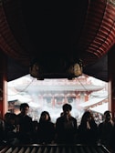 A group of people stand in a dimly lit interior space, silhouetted against bright daylight filtering through the entrance of a traditional temple. The foreground features individuals with hands clasped in prayer, while the background reveals the temple's intricate architecture and a bustling crowd outside, shrouded in a light mist or smoke.