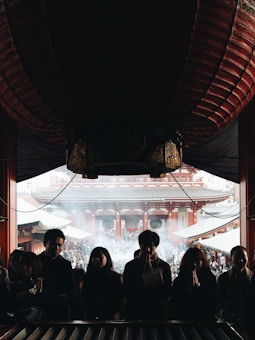 A group of people stand in a dimly lit interior space, silhouetted against bright daylight filtering through the entrance of a traditional temple. The foreground features individuals with hands clasped in prayer, while the background reveals the temple's intricate architecture and a bustling crowd outside, shrouded in a light mist or smoke.