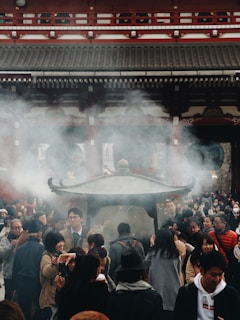 A bustling crowd gathers around a large incense burner, with smoke wafting into the air. The setting appears to be a traditional temple with ornate roofing and architectural details. People of various ages are engaged in conversations and activities, creating a lively atmosphere.