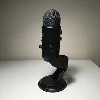 A black microphone is placed on a white surface, with its stand visible and several control knobs on the side. The lighting creates a soft shadow, giving the mic a sleek and modern appearance.