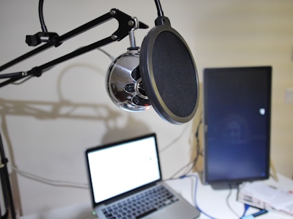 A microphone mounted on an adjustable arm is in focus, with a pop filter in front. Below it, a laptop with a bright screen and a flat-screen monitor are on a desk. Cables and a few papers are also visible on the desk, suggesting a recording or streaming setup.