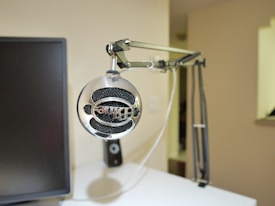 A modern desktop setup with a microphone mounted on an adjustable arm. The microphone is silver with a retro design, prominently displayed in the foreground. To the left, a monitor is partially visible. The background consists of a soft, neutral-colored wall.
