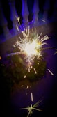 A sparkler emits bright, colorful sparks in a dark setting. The sparks create a vivid display of light against a background featuring a balustrade, with greenery faintly visible.