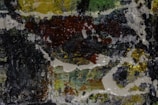 A textured abstract painting with a mixture of dark and vibrant colors. Thick brushstrokes create a dynamic pattern with a glossy finish.