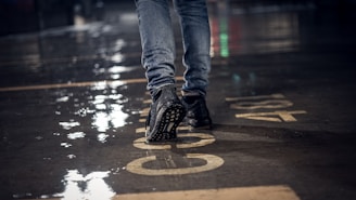 A close-up of a person walking on a wet concrete surface, wearing jeans and black sneakers. The ground reflects light due to the rain, and there are large numbers painted on it.