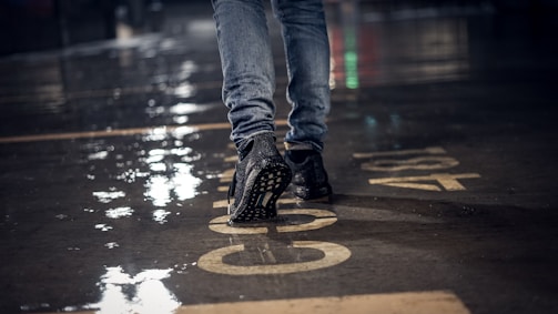 A close-up of a person walking on a wet concrete surface, wearing jeans and black sneakers. The ground reflects light due to the rain, and there are large numbers painted on it.