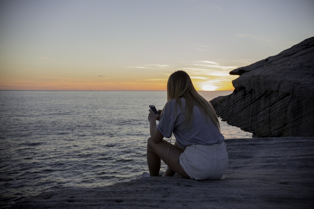 woman in grey shirts and white sorts sits on rock cliff by the beach