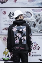 A person wearing a black hoodie with floral patterns and text stands in front of a wall with food advertisements and Chinese characters. The hoodie features cherry blossoms and a question addressing global warming. The person wears a white cap and sunglasses, and is turned away from the camera.
