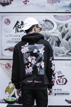 A person wearing a black hoodie with floral patterns and text stands in front of a wall with food advertisements and Chinese characters. The hoodie features cherry blossoms and a question addressing global warming. The person wears a white cap and sunglasses, and is turned away from the camera.