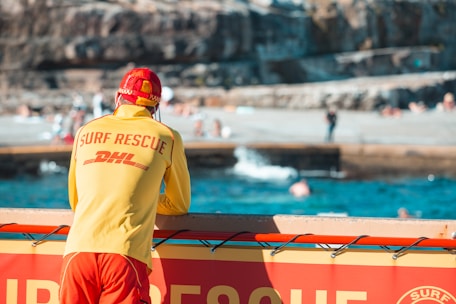 A lifeguard wearing a red and yellow surf rescue uniform stands by the seaside, overlooking the ocean. The setting is a beach with people swimming and relaxing in the background. Cliffs and a concrete area are visible further back.