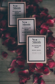 Three boxes of handmade soap labeled 'Rose Basil' from Vya Naturals are arranged on a surface surrounded by scattered red rose petals. The boxes are neatly aligned, displaying their minimalist design with a focus on natural ingredients.