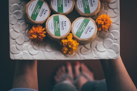 Handmade soap bars wrapped in paper are arranged on a textured, circular-patterned white dish along with vibrant orange marigold flowers. The dish appears to be held by two hands, with feet slightly visible in the background, suggesting a rustic or homemade setting.