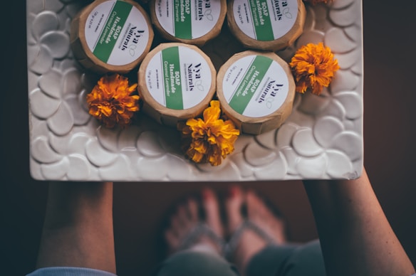 Handmade soap bars wrapped in paper are arranged on a textured, circular-patterned white dish along with vibrant orange marigold flowers. The dish appears to be held by two hands, with feet slightly visible in the background, suggesting a rustic or homemade setting.