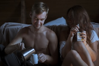 A shirtless man is pouring a drink from a stainless steel thermos into a white mug, sitting next to a woman with long hair who is smiling and holding a container. They are sitting close together in a cozy, dimly lit environment, possibly a bedroom or living area.